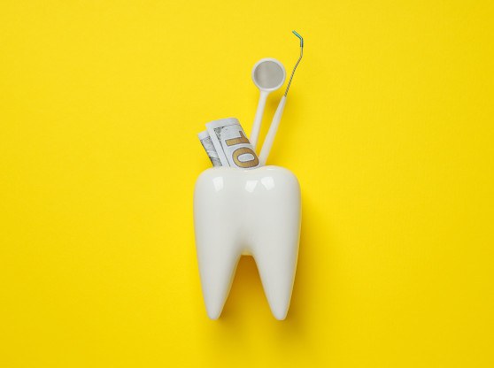 Large model tooth holding a $100 dollar bill and dental instruments with a yellow background